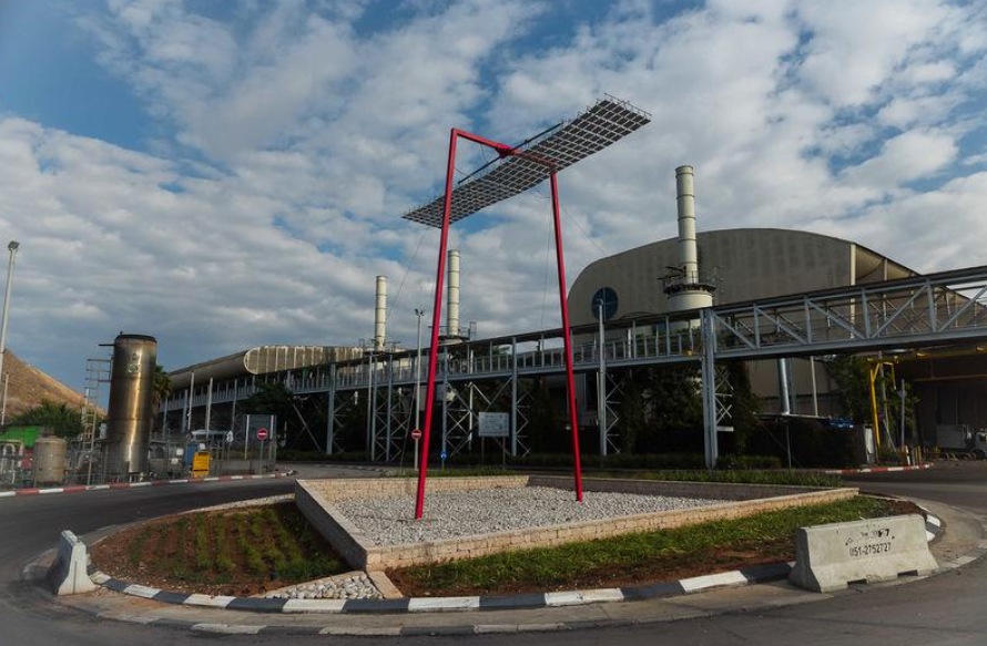 מבנה אדום מעל גינת אבנים מול מתקן תעשייתי וצריחים